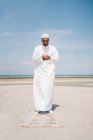 Garçon complet mâle islamique en vêtements blancs traditionnels debout sur le tapis et priant contre le ciel bleu sur la plage — Photo de stock