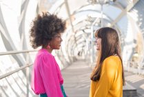 Side view of glad multiethnic LGBT couple of women holding hands while standing on bridge in city in sunny day and looking at each other — Stock Photo