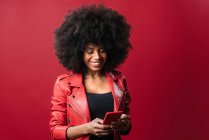 Eccitato afro americano femminile con acconciatura afro navigazione cellulare su sfondo rosso in studio — Foto stock