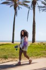 Full body side view of female with skateboard in hand strolling on sidewalk along tall palms against coast and sea — Stock Photo