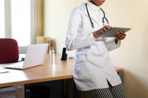 Обрезать молодую черную женщину-врача в медицинском халате со стетоскопом, работая с планшетом в современном офисе клиники — стоковое фото