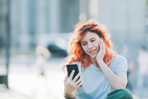 Seitenansicht fröhliche rothaarige Frau sitzt auf der Straße und Nachrichten in den sozialen Medien auf dem Handy — Stockfoto