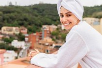 Mujer joven optimista en albornoz y toalla sonriendo y mirando a la cámara mientras se relaja en el balcón durante la rutina de cuidado de la piel en fin de semana - foto de stock