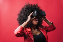 Вражена афро-американська жінка з відкритим ротом і в VR окуляри переживають віртуальну реальність на червоному тлі в студії — стокове фото