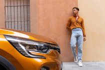 Jovem elegante étnico encaracolado cara na moda roupa encostado contra a parede perto estacionado moderno laranja automóvel na rua urbana — Fotografia de Stock