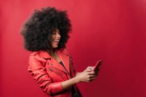 Excited African American female with Afro hairstyle browsing mobile phone on red background in studio — Stock Photo