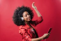 Délicieuse femme afro-américaine écoutant de la musique dans un casque et utilisant un téléphone portable tout en dansant sur fond rouge en studio — Photo de stock
