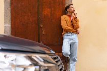 Jovem elegante etnia encaracolado cara na moda roupa encostada contra a parede perto estacionado automóvel moderno na rua urbana — Fotografia de Stock
