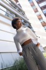 De baixo elegante auto-assegurada mulher afro-americana com as mãos no bolso olhando para longe, enquanto está perto de edifícios de apartamentos modernos no dia ensolarado na cidade — Fotografia de Stock