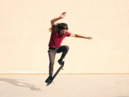 Homem skatista com cabelo ondulado realizando truque no skate enquanto salta sobre passarela e olhando para baixo no dia ensolarado — Fotografia de Stock