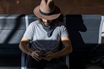 O cara de chapéu no aeroporto na sala de espera sentado esperando por seu voo, com fones de ouvido sem fio para ouvir música enquanto conversa com seu telefone inteligente, vista superior — Fotografia de Stock