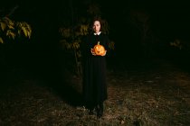 Female in black dress standing with glowing pumpkin lantern in dark woods on Halloween and looking at camera — Stock Photo