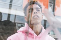 Através de vidro de sério jovem etnia hipster cara com penteado afro vestido com capuz rosa na janela e olhando para a câmera — Fotografia de Stock