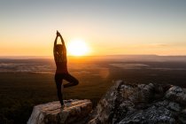 Молодая женщина йога практикует йогу на скале в горах со светом восхода солнца, вид сзади с одной ногой и поднятыми руками — стоковое фото