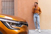 Young stylish ethnic curly haired guy in trendy outfit using smartphone while leaning against wall near parked modern orange automobile on urban street — Stock Photo