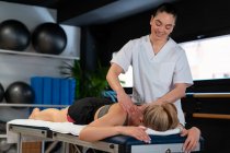 Friendly masseuse smiling and massaging shoulders of woman while working in physiotherapy clinic — Stock Photo