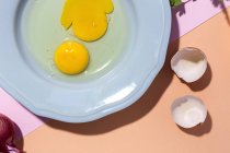 Vista aérea de huevos crudos en plato contra cáscaras de huevo y ramitas de perejil fresco sobre fondo de dos colores - foto de stock