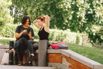 Щаслива молода різноманітна жінка найкращі друзі в активному одязі посміхаючись і ділячись мобільним телефоном, сидячи на кам'яній межі перед тренуванням йоги на відкритому повітрі в зеленому парку — стокове фото