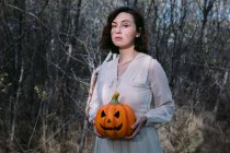 Donna in abito bianco in piedi con lanterna di zucca incandescente nel bosco di Halloween e guardando la fotocamera — Foto stock