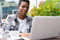 Happy African American male freelancer browsing and working remotely on laptop in outdoors cafe while sitting looking at camera at table with cup of coffee — Stock Photo