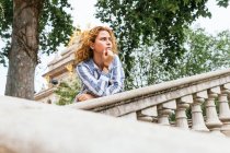 Low angle of thoughtful trendy male with long hair standing on staircase in street and leaning on hand while looking away — Stock Photo