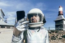 Alter Mann in Raumanzug und Helm surft Daten auf Smartphone, während er in der Nähe von Industriegebäuden mit raketenförmigen Antennen steht — Stockfoto