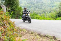 Full body of focused bearded ethnic male biker in black leather jacket and helmet riding modern motorbike on asphalt road amidst lush green trees growing in mountainous valley — Stock Photo