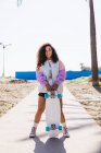Corpo pieno di femmina con skateboard in mano guardando la fotocamera sul marciapiede lungo palme alte contro costa e mare — Foto stock