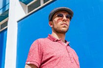 Junger bärtiger Mann mit lässigem roten Poloshirt und Mütze steht an einem Sommertag auf der Straße gegen eine blaue Wand — Stockfoto