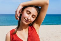 Cheerful female in summer clothes with pigtails standing with eyes closed on sandy shore with calm blue sea on sunny day — Stock Photo