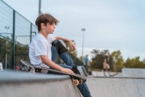 Vista lateral del adolescente sentado con monopatín en rampa en el parque de skate y mirando hacia otro lado - foto de stock