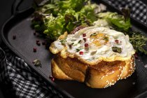 Fried egg on brioche served on tray with fresh lettuce for appetizing breakfast on black background — Stock Photo