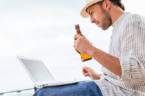 De baixo vista lateral do jovem blogueiro masculino em elegante desgaste e chapéu sentado com garrafa de cerveja na praia de areia e digitando no laptop durante as férias de verão na costa — Fotografia de Stock