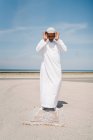 Ragazzo pieno maschio islamico in abiti bianchi tradizionali in piedi sul tappeto e pregando contro il cielo blu sulla spiaggia — Foto stock