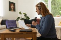 Delighted female radio host recording podcast while using mic and reading notes from paper at home — Stock Photo