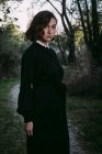 Donna senza emozioni che indossa un lungo abito nero in piedi nei boschi autunnali durante la celebrazione di Halloween guardando la fotocamera — Foto stock