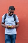 Афроамериканський чоловік з рюкзаком і мобільним телефоном посміхається, коли спирається на стіну — стокове фото