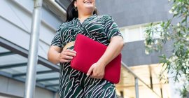 Знизу анонімно весела молода жінка в стильному смугастому вбранні з червоним чохлом ноутбука в руках, стоячи проти сучасної міської будівлі — стокове фото