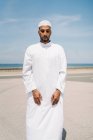 Ragazzo pieno maschio islamico in abiti bianchi tradizionali in piedi sul tappeto e pregando contro il cielo blu sulla spiaggia — Foto stock
