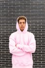 Confident serious young curly haired hipster guy in pink hoodie looking at camera against brick wall — Stock Photo