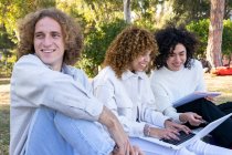 Multiethnische Männer und Frauen mit lockigem Haar sitzen mit Laptop und Notizblock auf dem Rasen im Park — Stockfoto