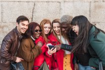 Compañía de amigos multiétnicos con estilo que se reúnen en la calle y ven videos divertidos en el teléfono móvil juntos - foto de stock