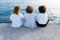 Back view of anonymous friends with curly hair sitting close against cityscape and embankment in sunlight — Stock Photo