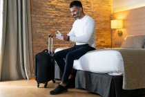 Angolo basso del viaggiatore maschio etnico positivo del raccolto seduto sul letto vicino alla valigia e al cellulare di navigazione in camera d'albergo — Foto stock