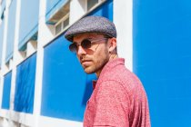 Junger bärtiger Mann mit lässigem roten Poloshirt und Mütze steht an einem Sommertag auf der Straße gegen eine blaue Wand — Stockfoto