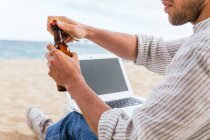 Side view of cropped unrecognizable male sitting with bottle of beer on sandy beach and typing on laptop during summer holidays on seashore — Stock Photo