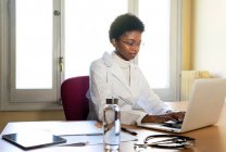 Jovem médico afro-americano datilografando relatório no laptop enquanto trabalhava na mesa no escritório da clínica moderna — Fotografia de Stock