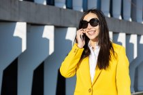 Улыбающаяся азиатская деловая женщина в желтом пальто и смартфоне идет по улице со зданием на заднем плане — стоковое фото