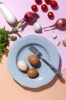 Vista superior de huevos de pollo en plato con tenedor contra ramitas de perejil fresco y tomates cherry sobre fondo de dos colores - foto de stock