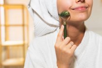 Colheita jovem fêmea com toalha na cabeça sorrindo e massageando o rosto com rolo de jade durante a rotina de cuidados com a pele em casa — Fotografia de Stock